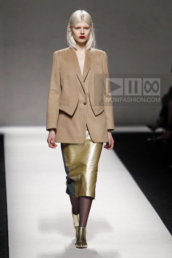 Model walking in a camel blazer, gold metallic midi skirt, and gold metallic ankle boots on a runway.