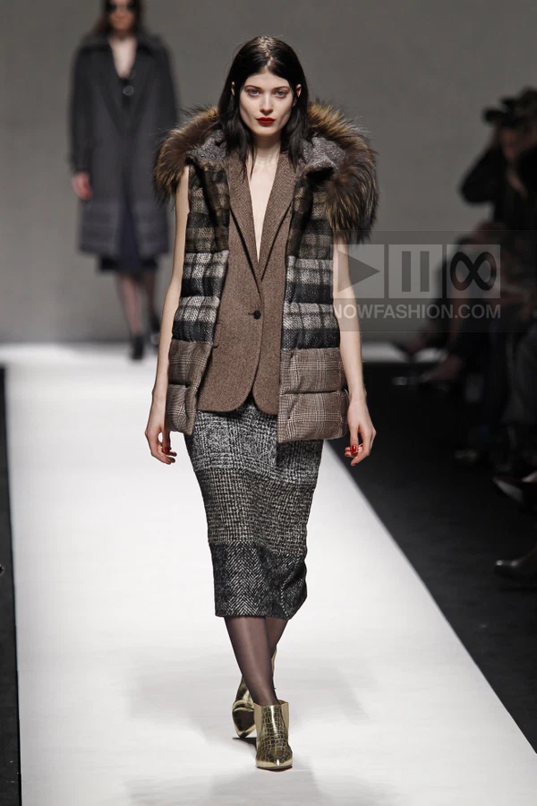 Model on runway wearing a brown blazer under a plaid puffer vest, paired with a gray tweed midi skirt and gold boots.