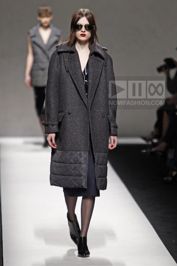 Model wearing a charcoal wool coat with a quilted lower panel over a dark dress and black ankle boots on a runway.
