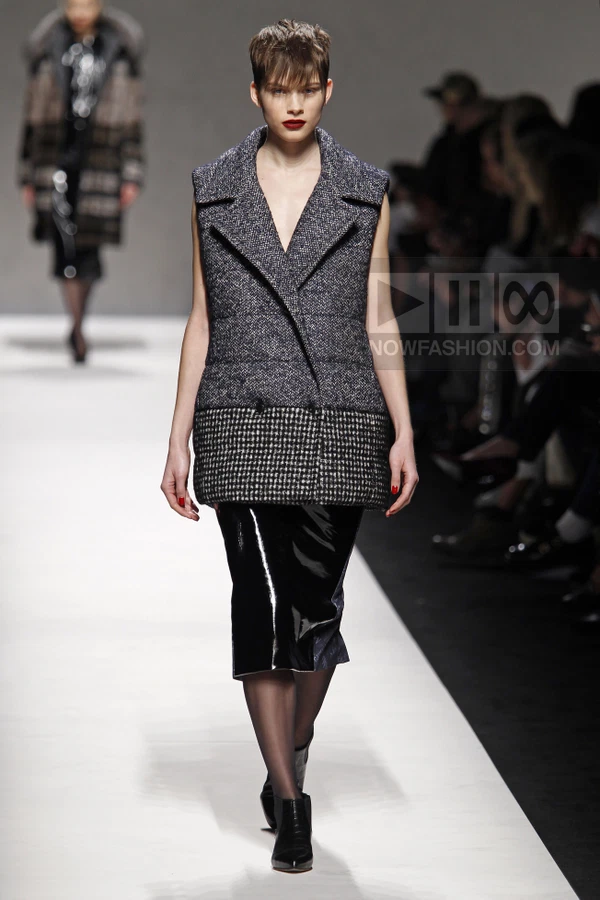 Model wearing a sleeveless double-breasted tweed vest over a black patent leather skirt on a runway.