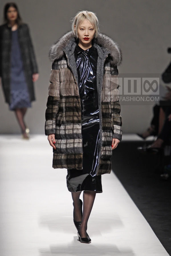 Model walking in a knee-length plaid puffer coat with fur hood over a black patent leather shirt and skirt on a runway.