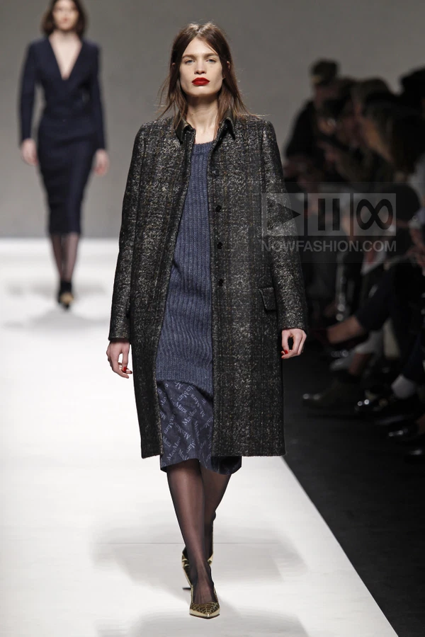 A model walks the runway wearing a dark textured wool coat over a ribbed blue knit dress and a printed navy skirt.