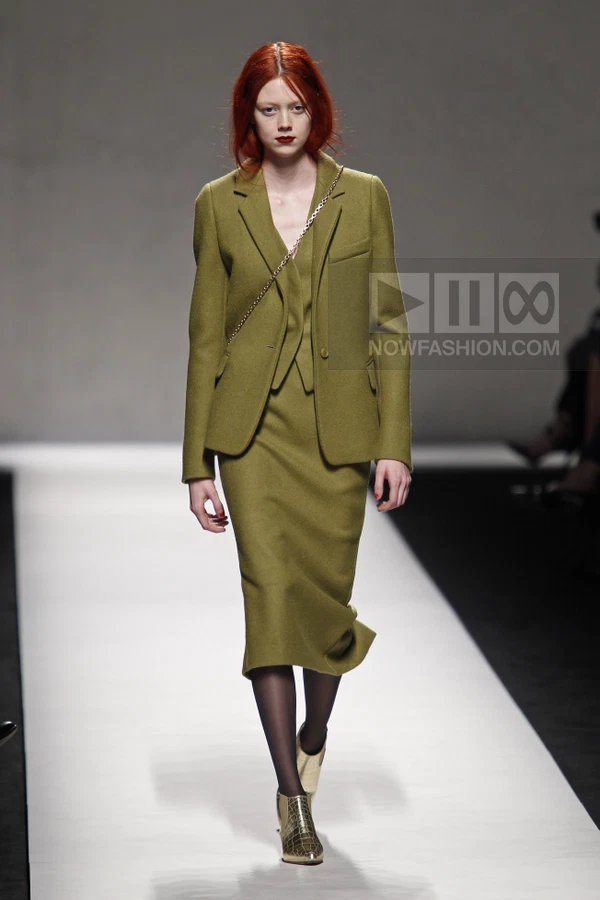 Model wearing an olive green wool blazer and matching midi skirt with a chain-strap bag on a runway.