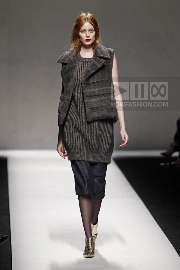 Model walking on a runway wearing a dark gray ribbed knit sweater dress and a matching quilted vest with ankle boots.