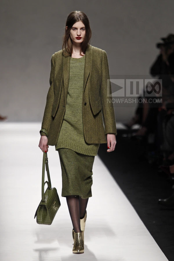 Model wearing an olive green wool blazer and matching knit dress with a green crocodile-embossed handbag on a runway.