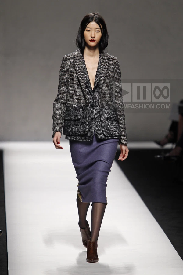 Model walking in a tweed blazer, purple midi skirt, and brown leather ankle boots on a runway.
