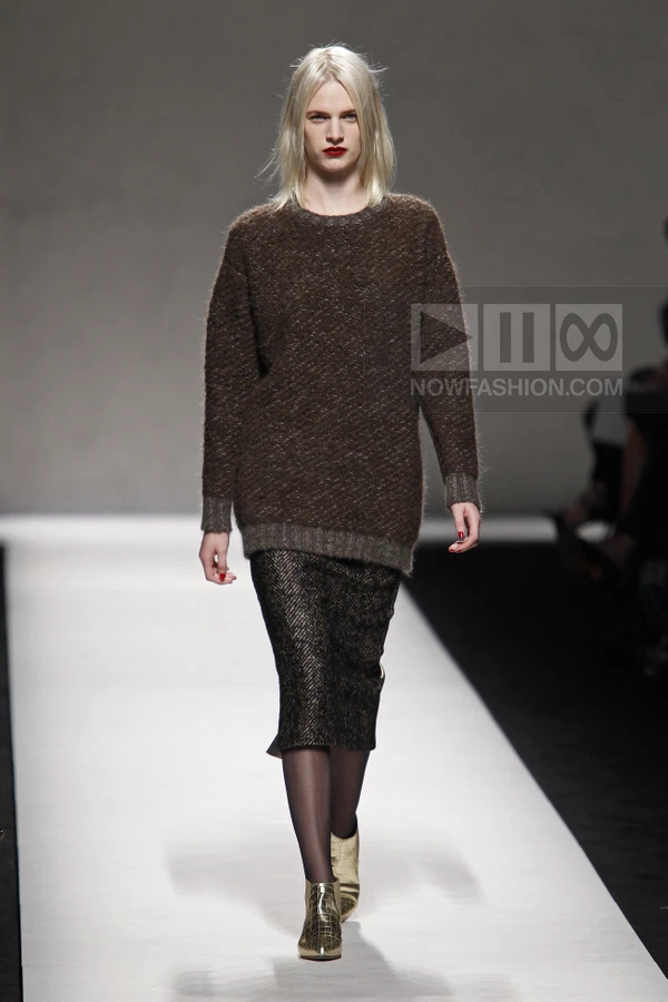 Model walking in a brown textured knit sweater and a black patterned midi skirt with gold ankle boots on a runway.