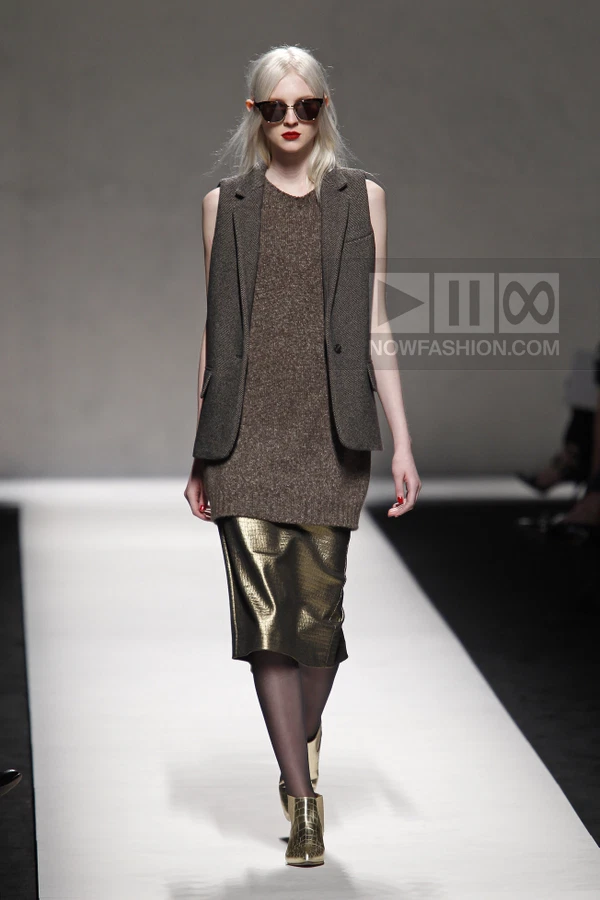 Model wearing a brown wool vest over a knit sweater dress and a metallic gold midi skirt on a runway.