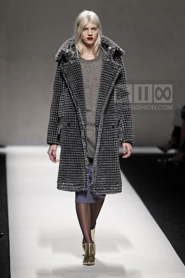 Model on runway wearing a gray houndstooth coat over a gray knit sweater and a purple skirt with gold ankle boots.