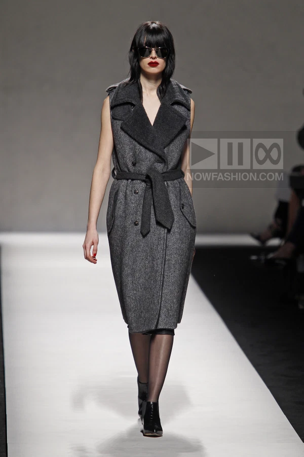Model wearing a sleeveless charcoal gray wool coat with a belted waist and dark lapels on a runway.