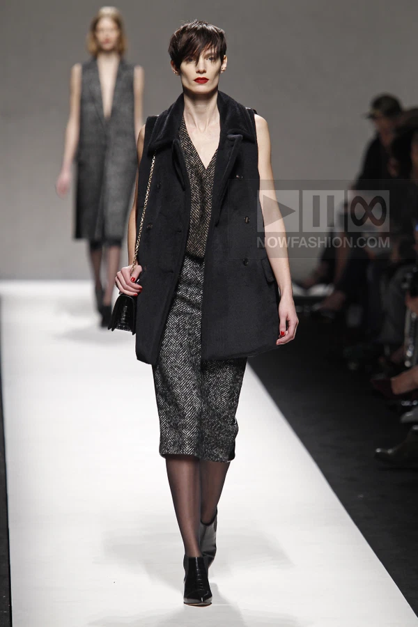 Model walking in a black sleeveless coat, herringbone midi skirt, and black ankle boots on a white runway.