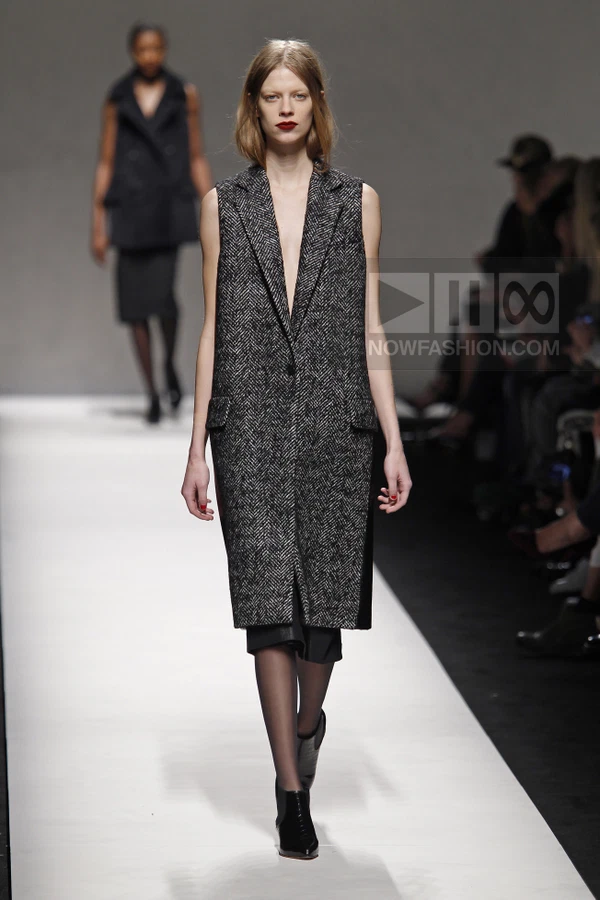 Model walking in a sleeveless black and white herringbone coat with a single button closure and flap pockets.