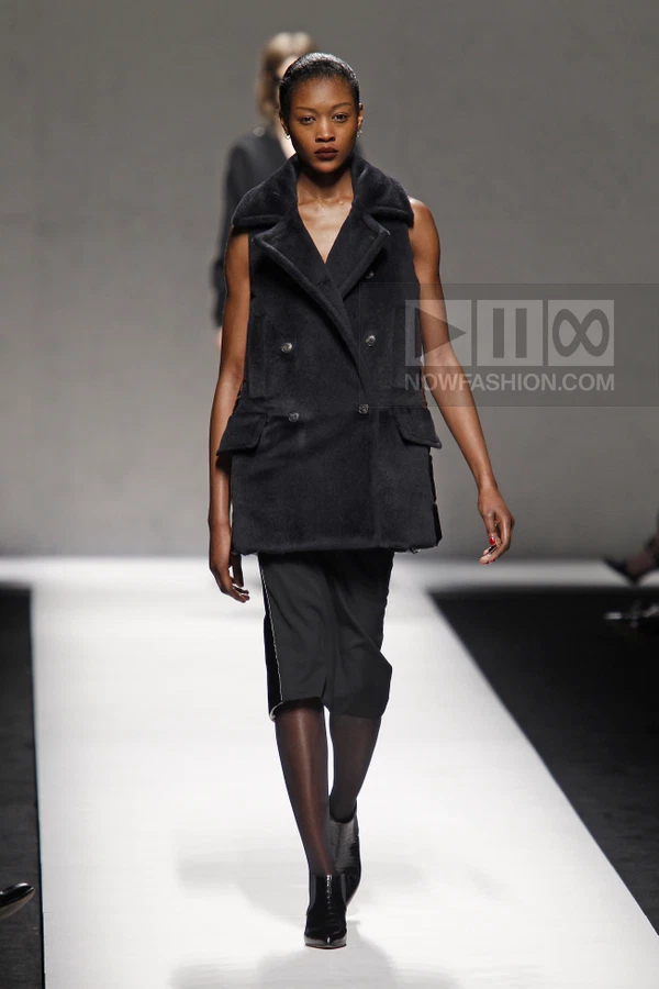 A model walks the runway in a black double-breasted sleeveless coat over a black knee-length skirt and dark tights.