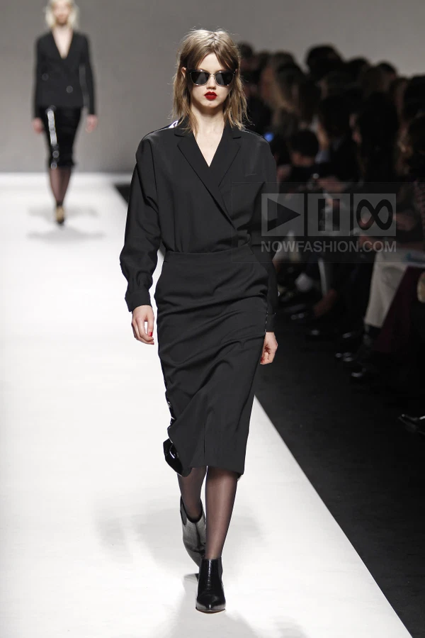 Model walking in a black wrap-style blouse and matching midi skirt on a runway.