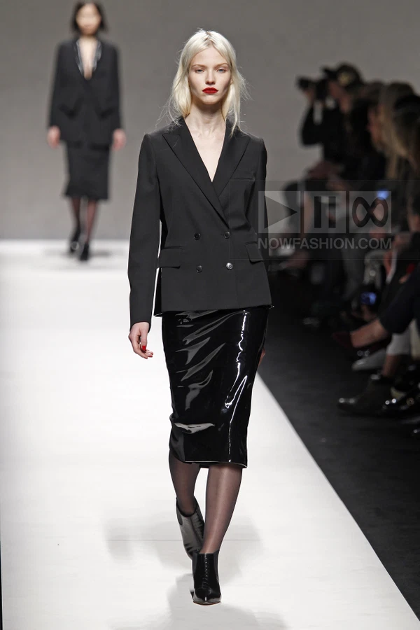 Model walking in a black double-breasted blazer and a black patent leather midi skirt on a white runway.