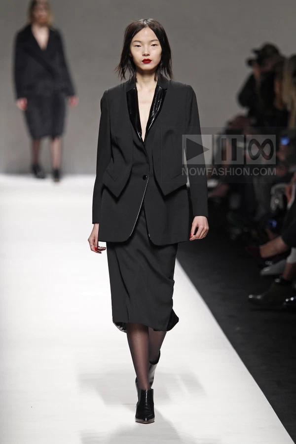 Model walking in a black blazer with patent lapels and a matching black midi skirt on a white runway.