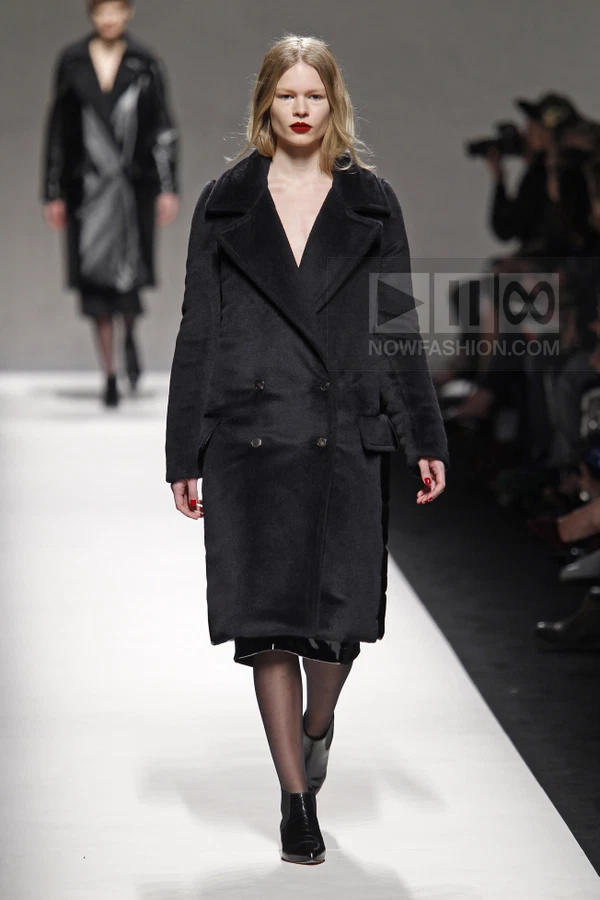Model walking in a black double-breasted wool coat over a black skirt, black tights, and black ankle boots on a runway.