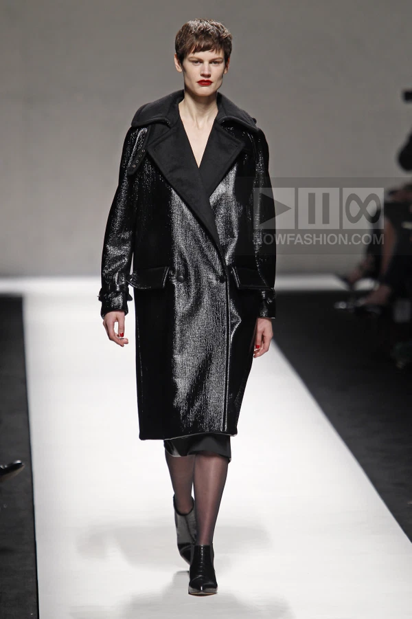 Model wearing a black patent leather coat with wide lapels and a dark skirt, walking on a white runway.