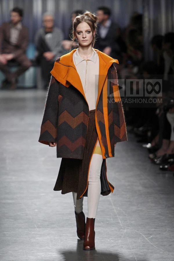 Model walking in a chevron-patterned wool coat with orange lining, worn over a cream top and brown skirt with ribbed tights.