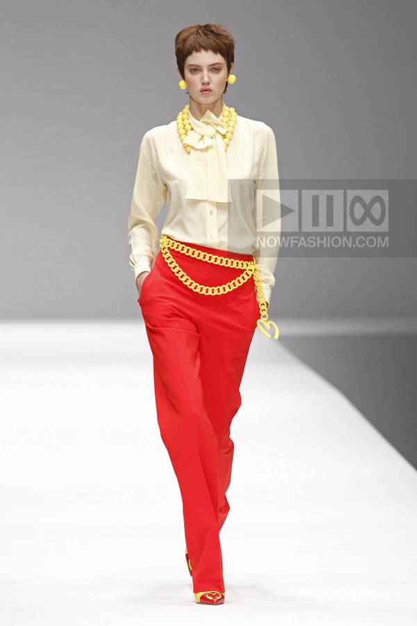 Model wearing a yellow striped blouse, red trousers, and yellow chain accessories on a runway.