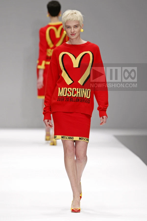 Model walking on a runway wearing a red knit sweater and matching skirt with yellow graphic logo and heart design.