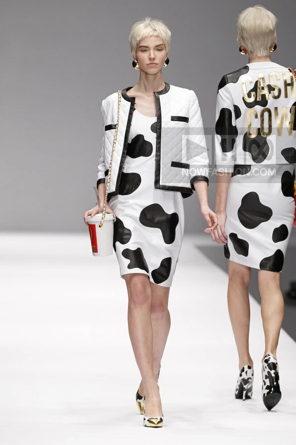 Model wearing a white and black cow-print dress and quilted jacket, carrying a cup-shaped bag on a runway.