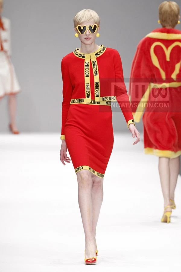 Model walks in a red knit skirt suit with yellow logo trim, heart-shaped sunglasses, and yellow earrings on a runway.