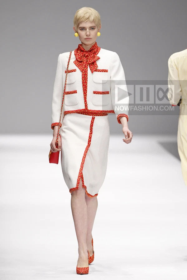 Model walking in a white skirt suit with red heart-patterned trim, a matching neck scarf, and a red chain-strap bag.