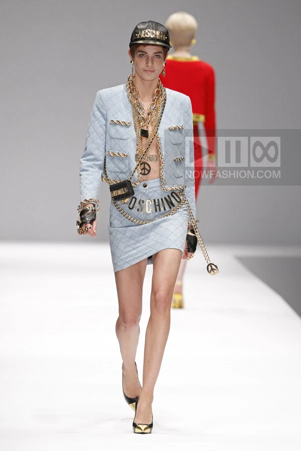 Model wearing a light blue quilted jacket and matching mini skirt with heavy gold chain accessories and a black cap.