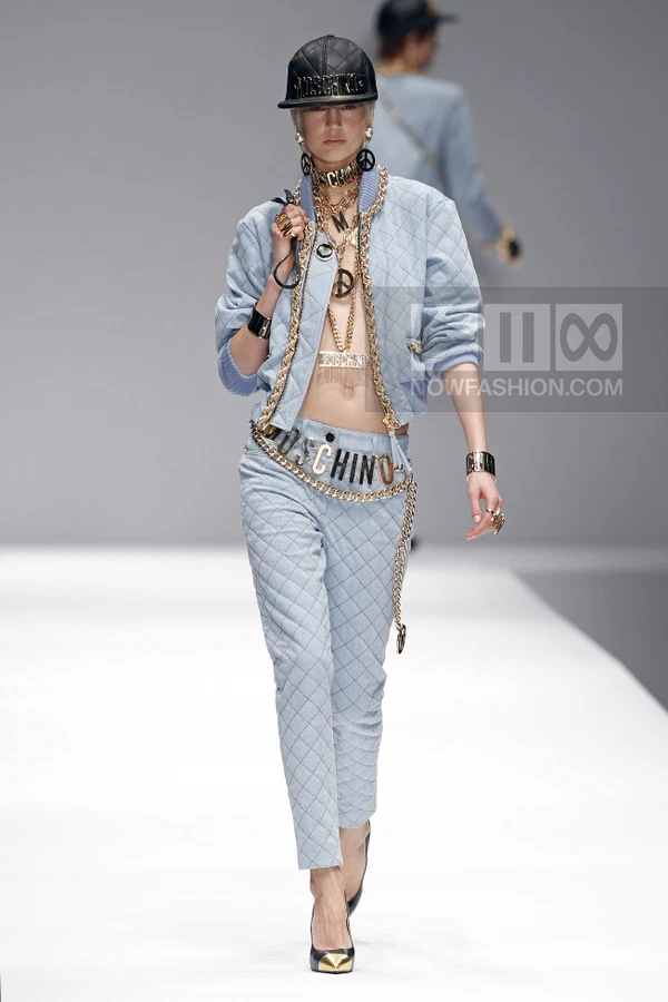 Model wearing a light blue quilted bomber jacket and matching quilted pants with heavy gold chain logo embellishments.