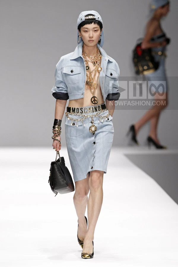 Model walking in a light-wash denim cropped jacket and matching button-front skirt with heavy gold chain accessories.