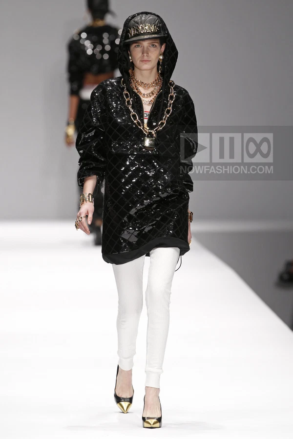 Model on runway wearing a black quilted hooded top, white leggings, and a baseball cap with gold chain accessories.