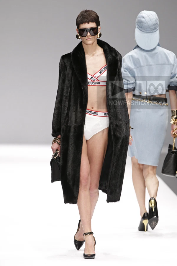 Model wearing a black fur coat over white logo-branded bra and briefs, walking on a runway with another model behind.