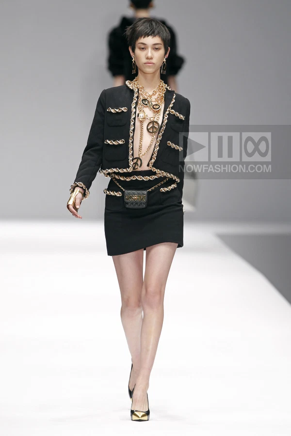 Model wearing a black cropped jacket with gold chain trim, matching mini skirt, and layered gold chain necklaces.