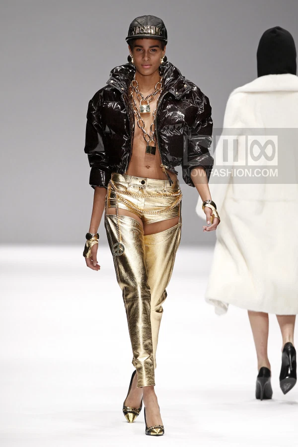 Model wearing a black puffer jacket, gold metallic chaps over shorts, and a baseball cap on a runway.