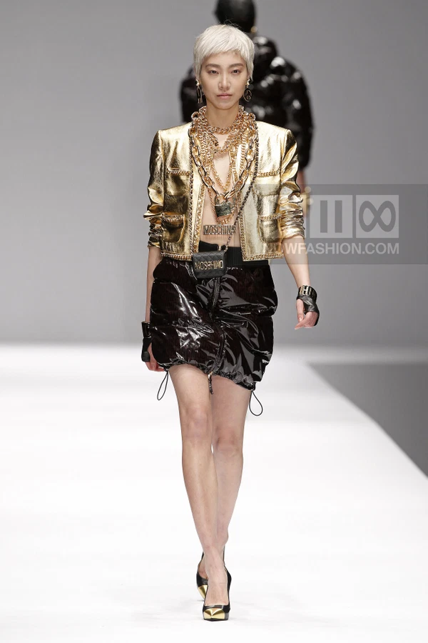Model wearing a metallic gold cropped jacket, black quilted mini skirt, and layered gold chain necklaces on a runway.