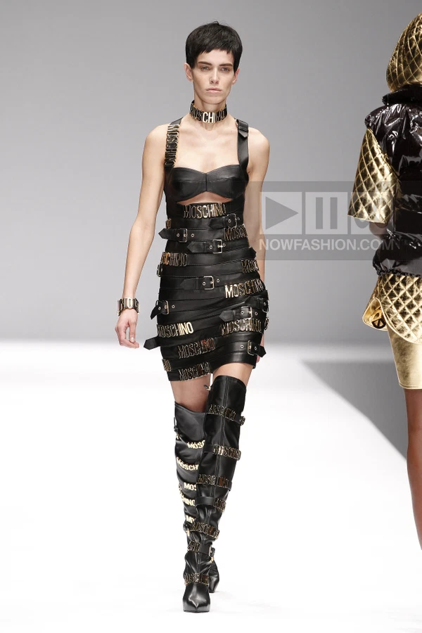 Model wearing a black leather mini dress covered in buckled straps and gold logo hardware, paired with matching…