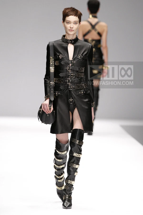 Model walking in a black leather coat with multiple buckled straps and gold logo hardware, paired with matching boots.