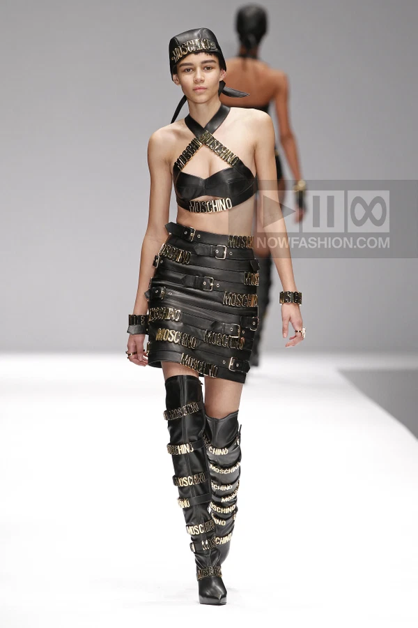 Model wearing a black leather halter top with gold logo hardware and a matching buckled leather mini skirt on a runway.