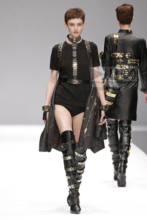 Model wearing a black logo-print t-shirt, leather vest, and thigh-high boots with gold logo hardware on a runway.