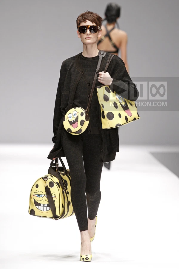 Model wearing a black monogram cardigan and leggings, carrying multiple yellow cartoon-print bags on a runway.