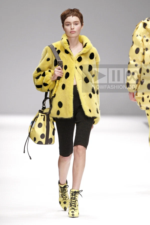 Model wearing a yellow polka-dot fur coat, black knit shorts, and yellow polka-dot lace-up boots on a runway.