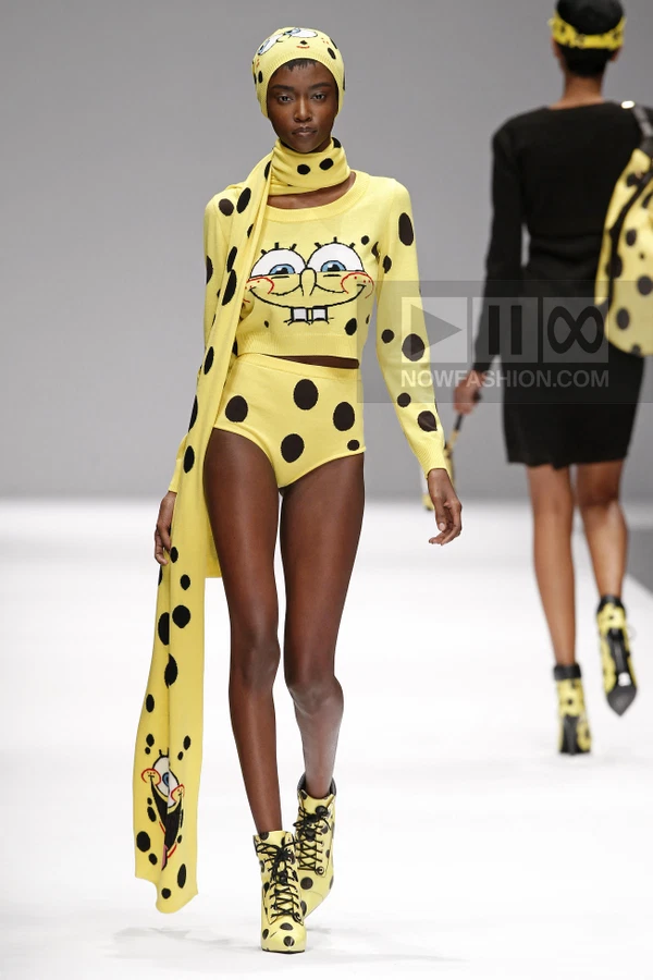 Model wearing a yellow knit crop top and high-waisted briefs with black polka dots and cartoon face graphic on runway.