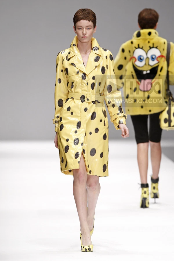 Model walking in a yellow trench coat with black polka dots, featuring a belted waist and pointed-toe pumps.