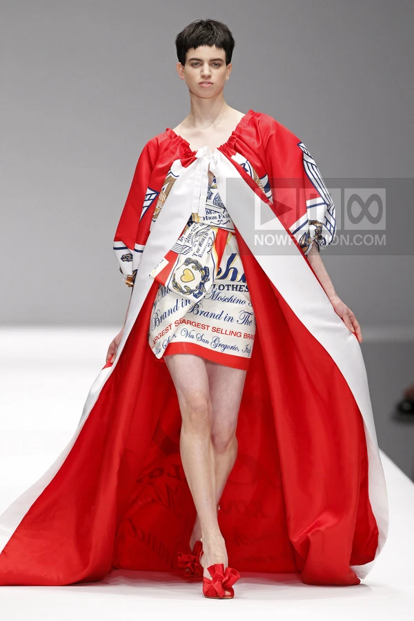 Model walking in a red and white oversized cape dress with graphic text prints and a mini skirt, wearing red heels.