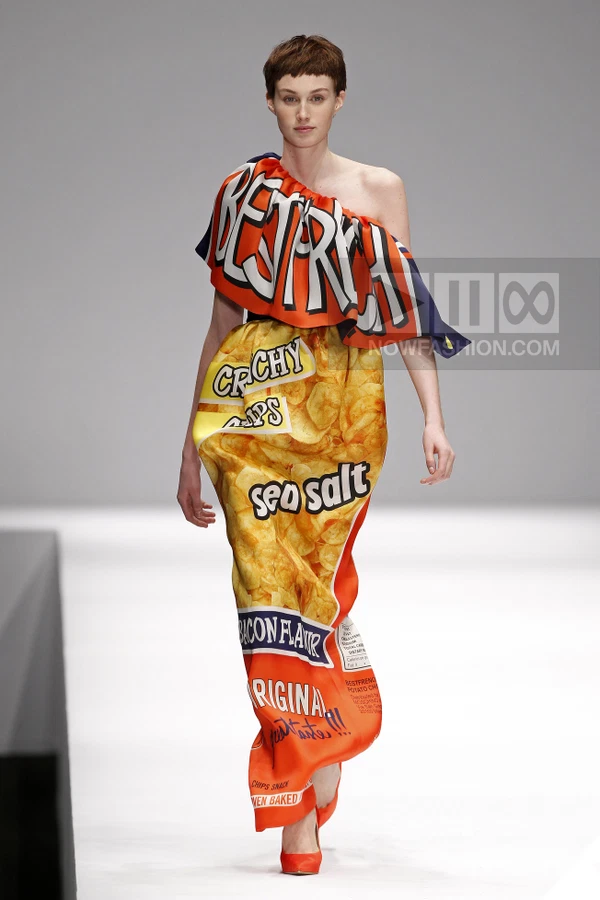 Model walking in a floor-length, one-shoulder dress printed with a potato chip bag design, featuring orange and red tones.