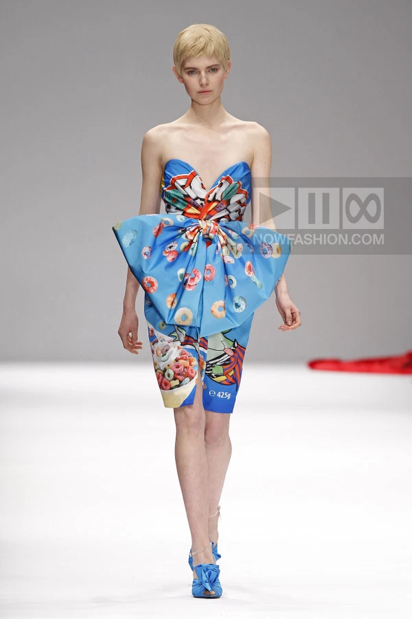 Model wearing a strapless blue midi dress with a large front bow and cereal box graphic print on a white runway.