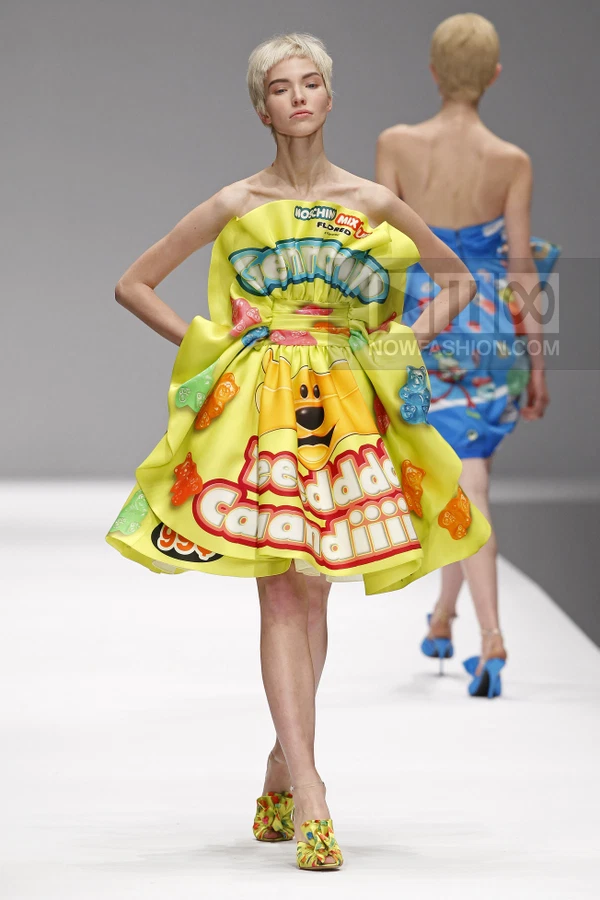Model wearing a yellow strapless mini dress with graphic candy prints and 3D gummy bear appliques on a runway.
