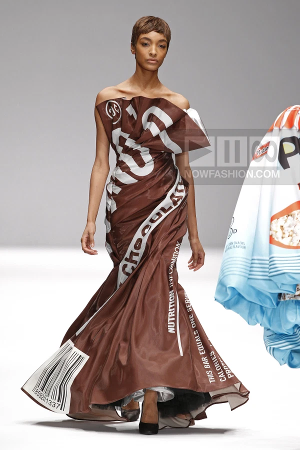 Model wearing a brown strapless gown printed with chocolate bar packaging graphics, including a barcode and text.