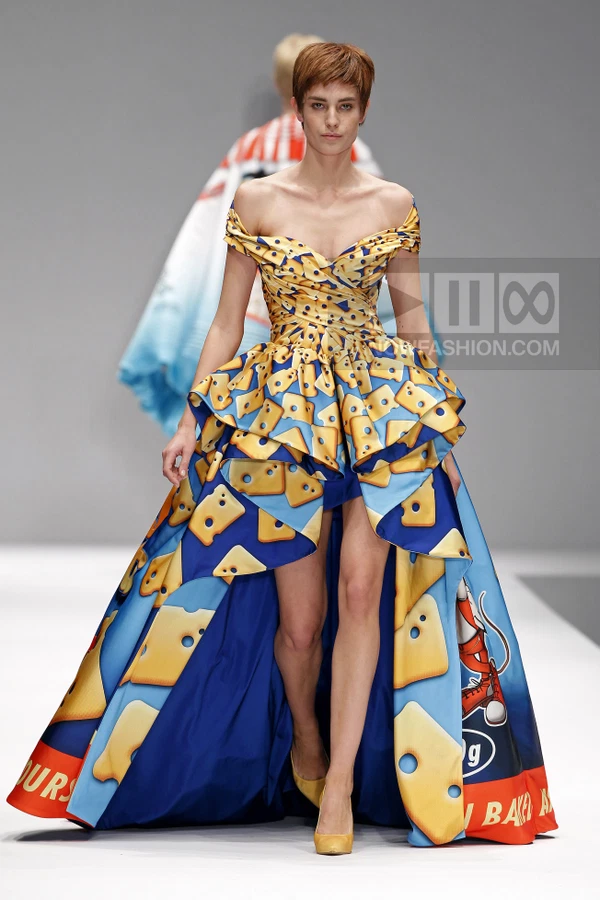 Model wearing a yellow and blue off-the-shoulder gown with a cheese print and high-low hem on a runway.